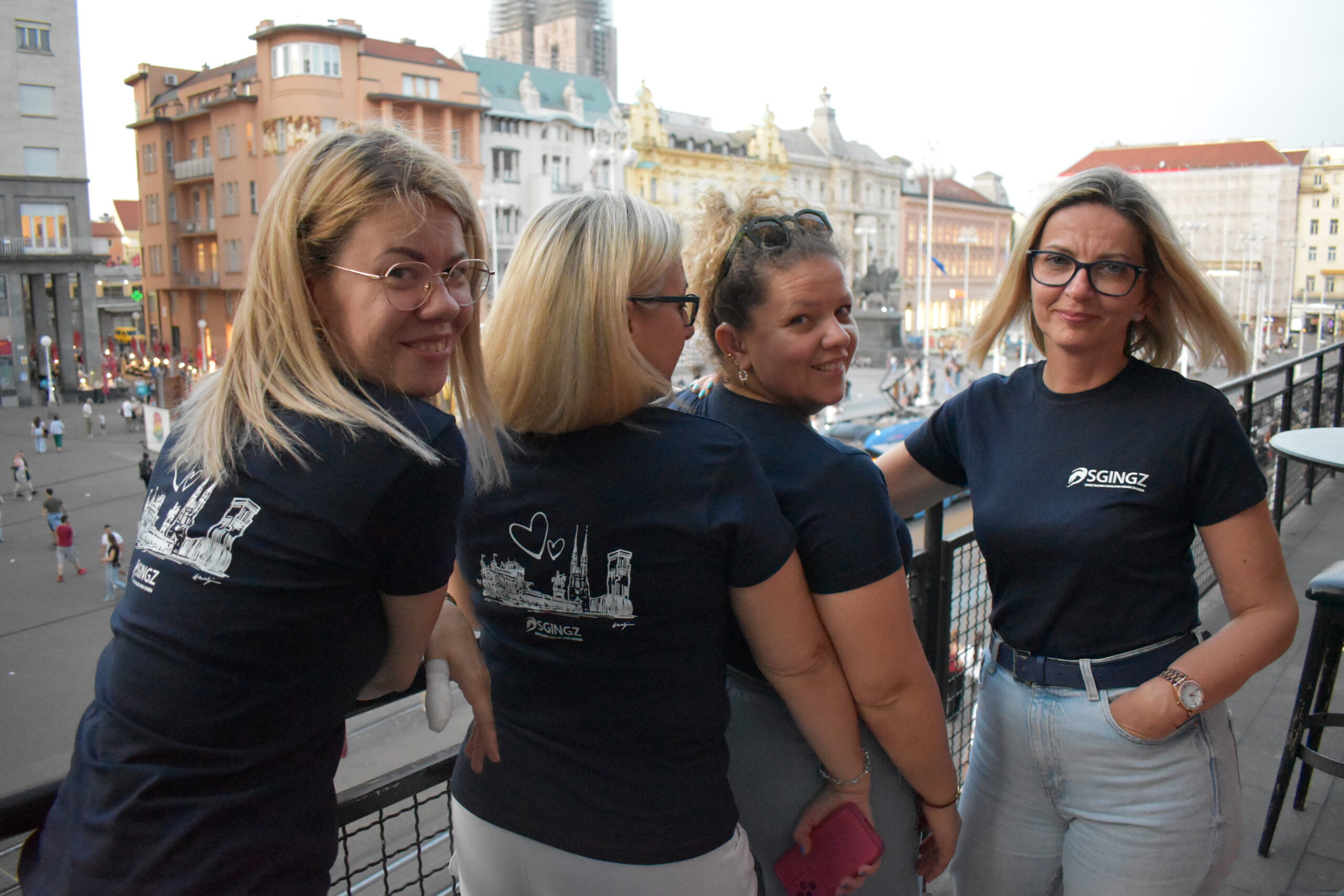 Na slici se nalaze četiri žene koje stoje na balkonu s pogledom na glavni zagrebački trg – Trg bana Jelačića. Sve nose tamnoplave majice s logotipom "SGINGZ", a na stražnjoj strani majica otisnuta je bijela ilustracija poznatih zagrebačkih građevina uz srce, što dodatno naglašava lokalni karakter i ponos. Tri žene okrenute su leđima prema kameri i nasmiješeno gledaju unatrag, dok je jedna (krajnje desno) okrenuta prema kameri, pozirajući s rukom u džepu. U pozadini se vidi gradska vreva, povijesne zgrade i prepoznatljiva arhitektura Zagreba, uključujući toranj katedrale koji je djelomično pod skelama. Atmosfera je opuštena i prijateljska, a fotografija odaje dojam timskog duha i zajedništva na događanju koje se očito održava u središtu Zagreba.