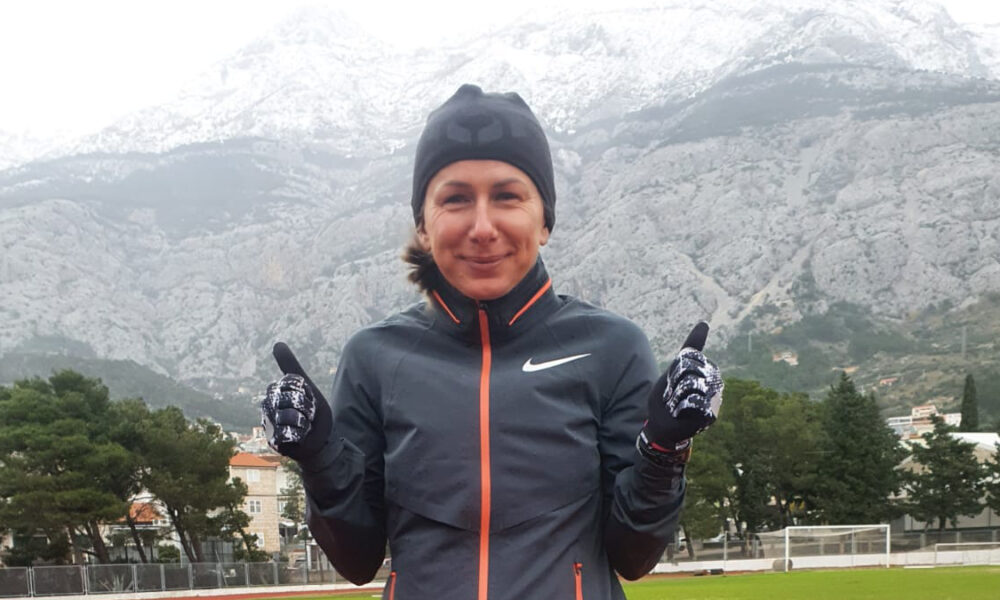Žena u sportskoj odjeći i rukavicama stoji nasmijana ispred snijegom prekrivenih planina. Nosi kapu i jaknu s Nike logotipom te pokazuje palčeve prema gore. U pozadini su borovi, sportsko igralište i planinski krajolik.