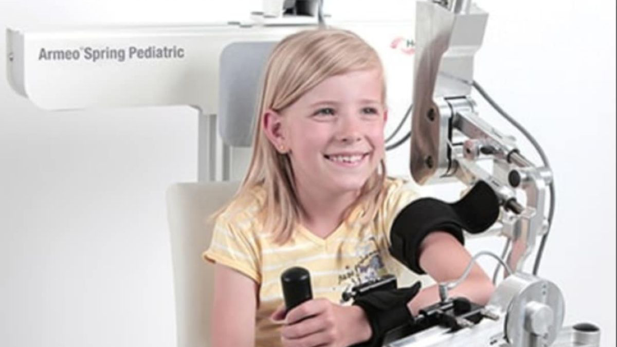 Na fotografiji je prikazano dijete koje sjedi i koristi rehabilitacijski uređaj za ruku nazvan "Armeo Spring Pediatric". Uređaj je dizajniran za pomoć djeci u terapiji oporavka gornjih ekstremiteta, posebno ruke. Dijete drži ručku koja je povezana s mehaničkim sustavom, a koji omogućuje vođene i precizne pokrete. Uređaj ima više povezanih dijelova koji podupiru ruku i omogućuju izvedbu terapijskih aktivnosti. Ovakvi uređaji koriste se u rehabilitacijskim centrima kako bi djeci s motoričkim poteškoćama omogućili lakši i učinkovitiji oporavak – često kroz igru i interaktivne zadatke.