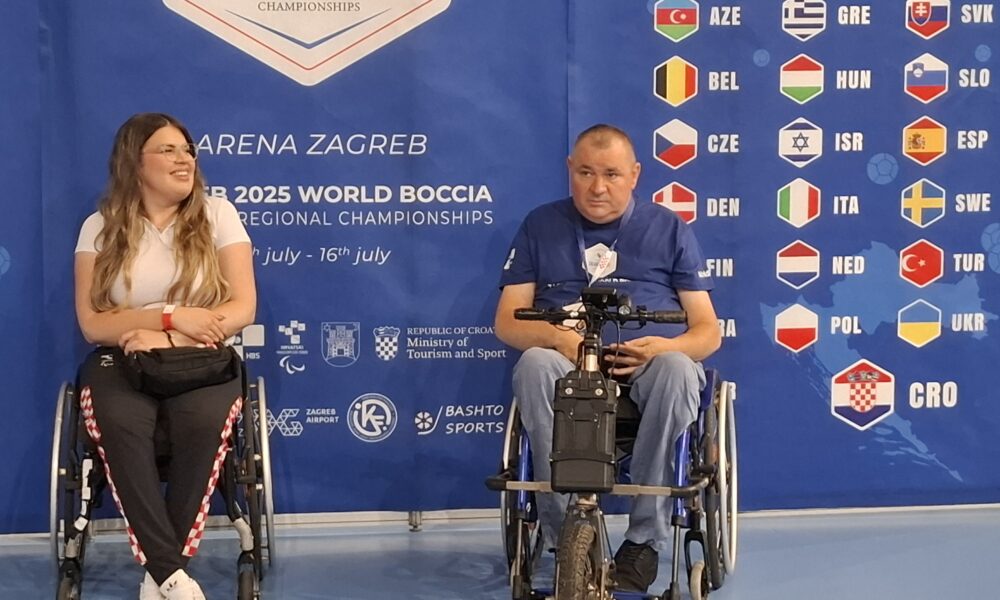 Fotografija prikazuje dvoje ljudi u invalidskim kolicima koji poziraju ispred plakata na kojem piše 'Europian World boccia chempionship'. Plakat je plave boje. Na njemu se također nalaze zastave svih zemalja koje će sudjelovati na natjecanju.