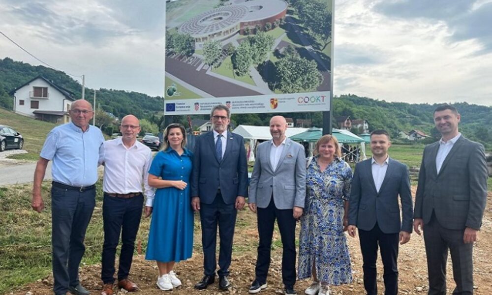 Na fotografiji se nalazi osam osoba koje stoje ispred informativne ploče s vizualizacijom budućeg Centra za odgoj i obrazovanje u Krapinskim Toplicama. Ploča prikazuje moderan kružni objekt okružen zelenilom, a na njoj su istaknuti logotipi Europske unije, Ministarstva znanosti, obrazovanja i mladih, Krapinsko-zagorske županije te samog Centra (COOKT). Osobe na slici su svečano odjevene i poziraju nasmiješene, što upućuje na važan događaj – vjerojatno obilježavanje početka radova na izgradnji Centra. U pozadini se vidi brežuljkasti krajolik s kućama, što je karakteristično za područje Krapinskih Toplica, kao i šatori koji sugeriraju organizirani događaj. Atmosfera fotografije je svečana i optimistična.