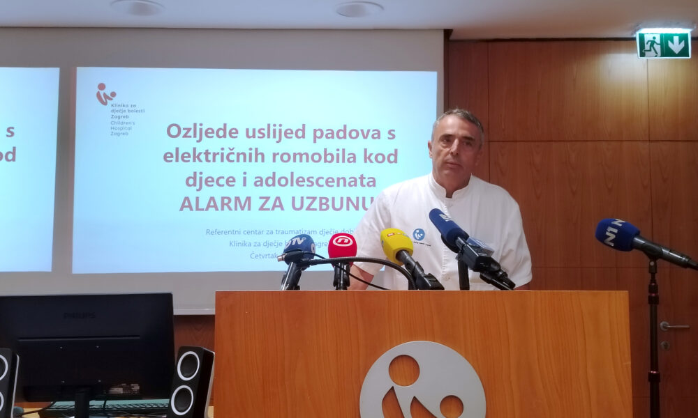 Liječnik u bijeloj kutiji stoji za govornicom ispred projekcije na kojoj piše: „Ozljede uslijed padova s električnih romobila kod djece i adolescenata – ALARM ZA UZBUNU!“ Na govornici i ispred nje su postavljeni brojni mikrofoni hrvatskih medija, a u pozadini se nalaze logotipi Kliničkog bolničkog centra i projekcija s podacima o prezentaciji.