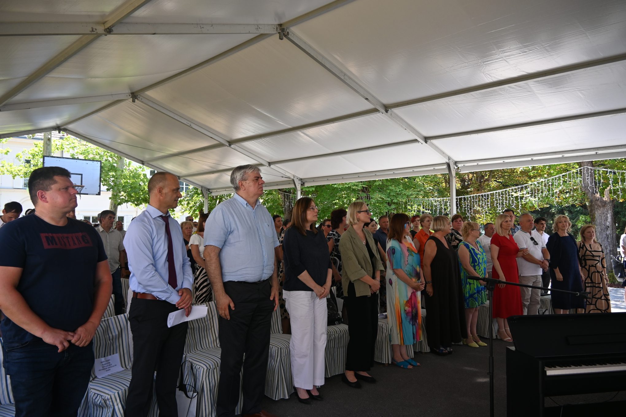 Na fotografiji je prikazan događaj na otvorenom, vjerojatno svečana prigoda poput proslave ili ceremonije: Grupa ljudi okupljena je ispod velikog bijelog šatora, a njihov stil odijevanja varira – neki su formalno odjeveni, dok su drugi u ležernijoj odjeći U pozadini se vidi zelenilo, drveće i decentne svjetlosne dekoracije koje stvaraju svečani i ugodan ugođaj Na desnoj strani fotografije nalazi se crni klavir, što sugerira mogućnost glazbenog dijela programa Cijeli prizor odiše toplinom i zajedništvom, kao da se svi zajedno raduju nečemu važnom ili radosnom.