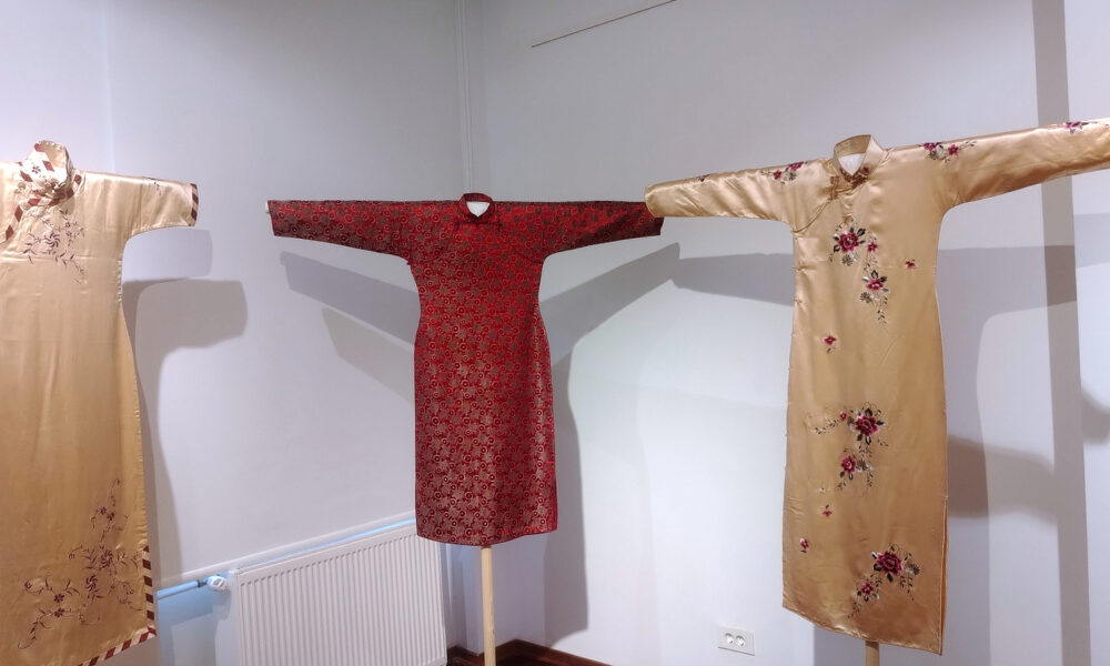 Tri tradicionalne kineske haljine qipao izložene na drvenim stalkovima u muzejskoj prostoriji. Haljine su dugih rukava i izrađene od svilenog materijala s bogatim uzorcima. Dvije su zlatne boje s cvjetnim vezom, dok je srednja tamnocrvena s geometrijskim uzorkom. Prostorija je jednostavno osvijetljena, s bijelim zidovima i drvenim podom.