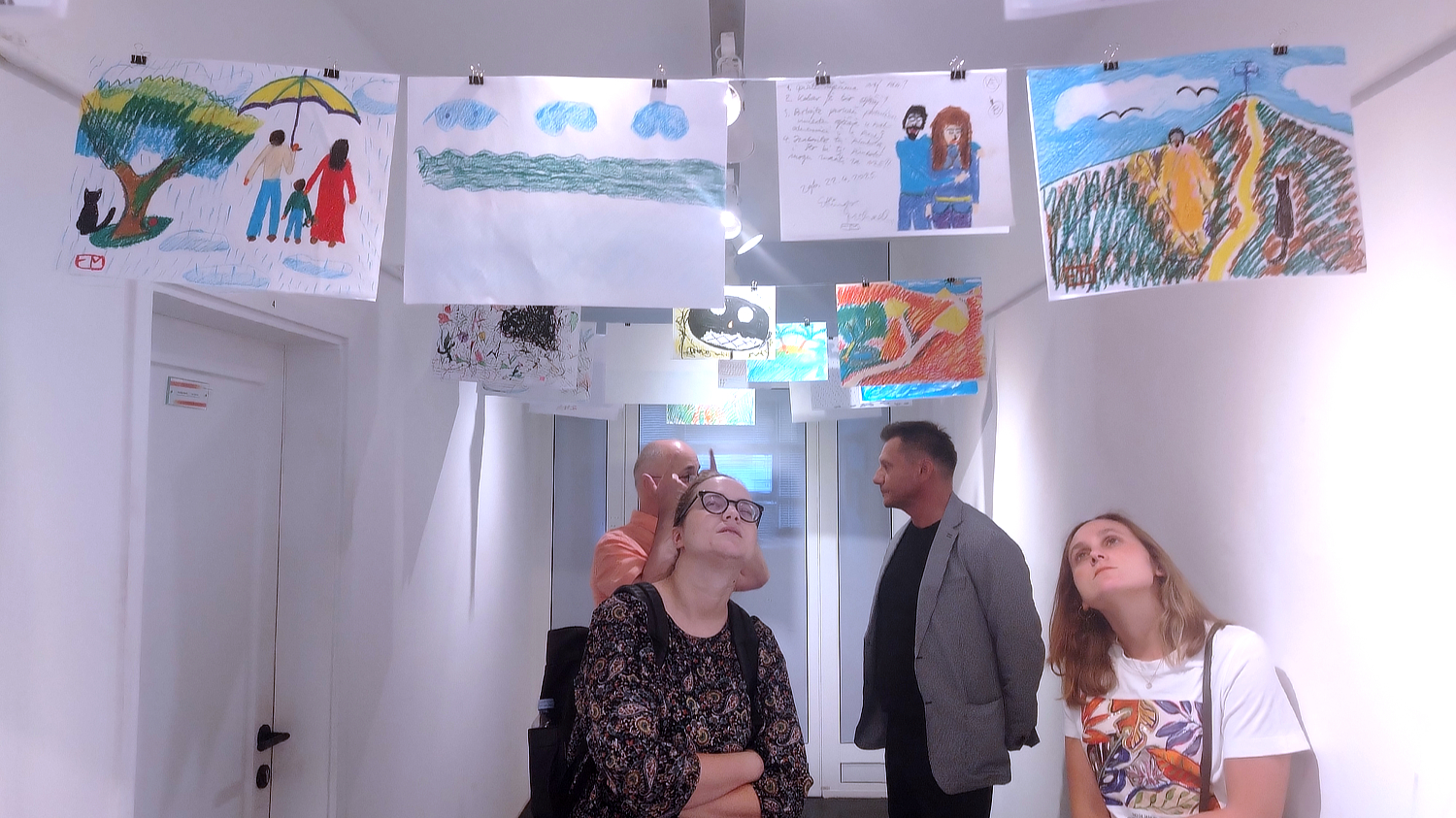 Unutrašnji prostor galerije s bijelim zidovima i stropnom rasvjetom, u kojem su dječji crteži obješeni na žici iznad glava posjetitelja. Crteži su raznobojni i prikazuju motive poput obitelji s kišobranom, pejzaža, planina i likova. U prostoru se nalaze četiri osobe koje promatraju izložene radove, dvije žene u prvom planu gledaju prema gore s izrazima znatiželje, dok se dva muškarca u pozadini razgovaraju.