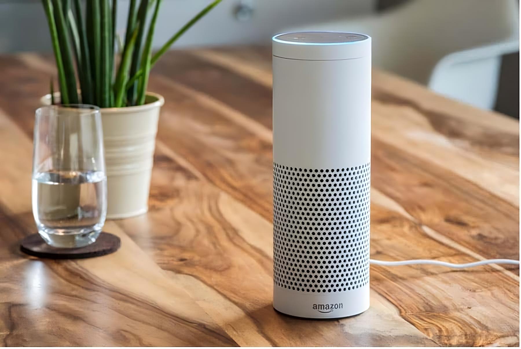 Pametni zvučnik Amazon Echo u bijeloj boji s plavim svjetlom na vrhu stoji na drvenom stolu. Pored njega se nalazi čaša vode na podlošku i bijela tegla s biljkom. U pozadini je zamućen interijer prostorije.