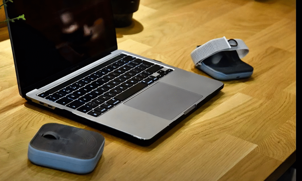 Prikaz prijenosnog računala (MacBook) na drvenom stolu, uz dva dodatna uređaja s remenima i plastičnim kućištem, koji izgledaju kao asistivne tehnologije. U pozadini je biljka u tegli. Ambijent djeluje moderno i tehnološki usmjeren.