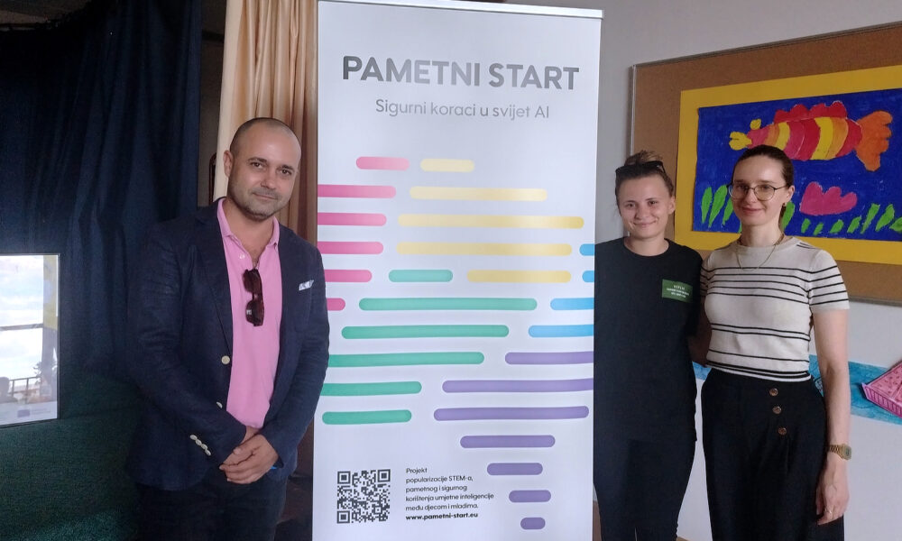 Tri osobe stoje nasmijane pored promotivnog roll-upa projekta „Pametni start – sigurni koraci u svijet AI“. Roll-up sadrži šareni grafički prikaz, slogan i QR kod. U pozadini se nalaze dječji crtež i školska oglasna ploča, što sugerira da se događanje odvija u edukativnom okruženju.
