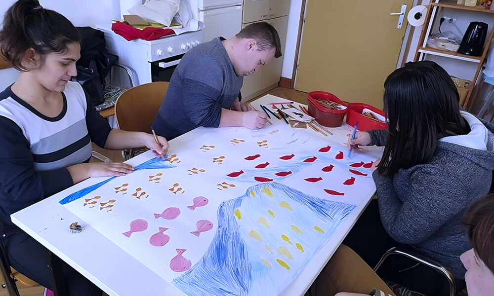 Četvero mladih osoba sjedi oko stola i sudjeluje u zajedničkoj likovnoj aktivnosti. Na bijelom papiru većih dimenzija crtaju i boje motive riba, valova i kapljica u raznim bojama. Atmosfera je opuštena i kreativna, a prostor je jednostavno uređen s kuhinjskim i radioničkim elementima. Aktivnost potiče razvoj kreativnosti i socijalnih vještina kod djece i mladih s teškoćama.