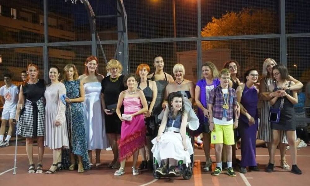 Na fotografiji se nalazi skupina ljudi koji poziraju na vanjskom košarkaškom terenu, pod večernjim nebom. Mnogi su odjeveni svečano — u elegantne haljine, odijela, košulje i kravate — što sugerira da je riječ o posebnoj prigodi, možda proslavi, svečanosti ili modnoj reviji. Jedna osoba sjedi u invalidskim kolicima, što fotografiji dodaje element inkluzivnosti i zajedništva.