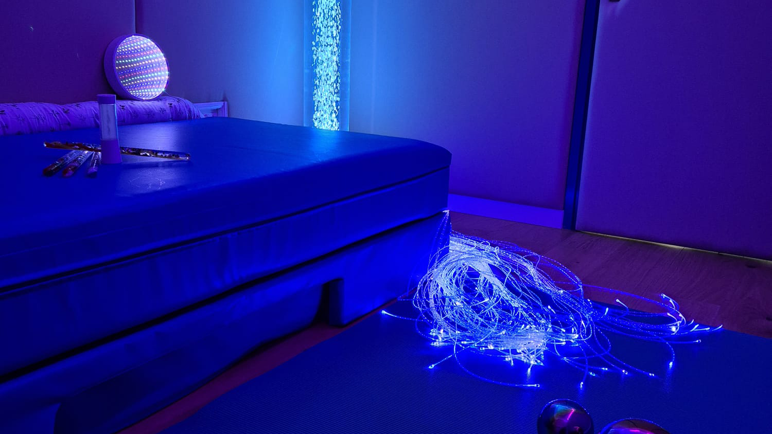 Prostor senzorne sobe u plavom ambijentalnom svjetlu. Na podu je plavi madrac, a pored njega svjetleća vlakna optičkih niti koje svijetle u plavim tonovima. U pozadini se nalazi stup s mjehurićima i svjetlom, dok su na madracu raspoređeni predmeti poput pješčanog sata, šarenih svjetlećih štapića i reflektirajuće lopte. Prostor djeluje umirujuće i osmišljen je za senzornu stimulaciju.