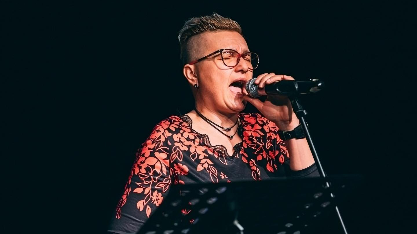 Pjevačica kratke kose s modernom frizurom i naočalama pjeva u mikrofon na pozornici. Odjevena je u crnu majicu s crveno-bijelim cvjetnim uzorkom. U ruci drži mikrofon, a ispred nje se vidi stalak s notama. Pozadina je potpuno tamna, fokus je na izvođačici.