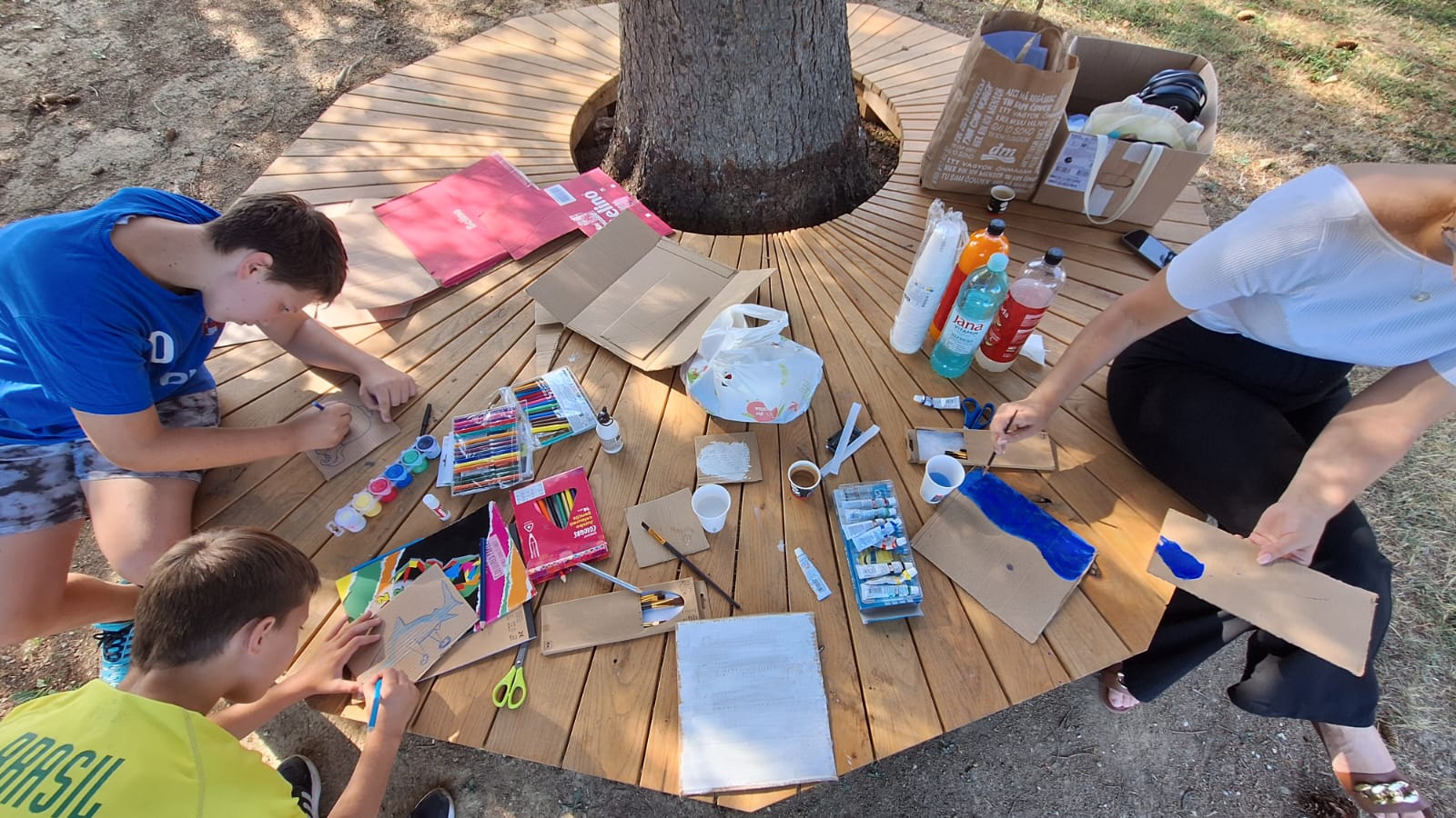 Na ovoj fotografiji vidi se šarmantna scena kreativne radionice na otvorenom. 🌳 Opis prizora: Drveni stol je smješten oko stabla, u hladu krošnje, što prostoru daje prirodan i opušten ambijent. Na stolu se nalaze razni umjetnički materijali: tempere, četkice, olovke, škare, ljepilo, karton, boce s vodom i sokom, šalice, papirnate vrećice, te kutije s bojicama i flomasterima. Tri osobe sudjeluju u aktivnostima – jedna slika plavom bojom, druga crta olovkom, a treća je usmjerena prema sredini stola. Papir s tekstom u donjem dijelu slike nažalost nije čitljiv, ali dodaje osjećaj da je u pitanju vođena radionica ili zadatak. ✨ Cijeli prizor zrači kreativnošću, zajedništvom i toplom ljetnom atmosferom. Ako si ti sudjelovala u ovome, djeluje kao predivno iskustvo.