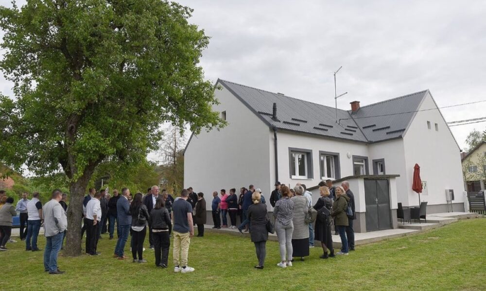 Na fotografiji se nalazi grupa ljudi okupljena ispred bijele kuće s tamnim krovom. Kuća ima nekoliko prozora i ulaznih vrata, a ispred nje se prostire travnjak s velikim drvetom koje stvara ugodan hlad. Ljudi su raspoređeni u manjim grupama — neki razgovaraju međusobno, dok drugi gledaju prema kući, što sugerira da se možda održava neki društveni događaj ili okupljanje. U pozadini se mogu vidjeti još nekoliko kuća i drveće, što daje dojam mirnog naselja. Cijela scena odiše opuštenom atmosferom, kao da se radi o proslavi, susretu s prijateljima ili čak obiteljskoj svečanosti.