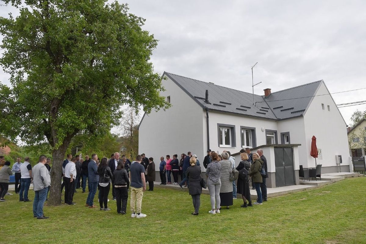 Na fotografiji se nalazi grupa ljudi okupljena ispred bijele kuće s tamnim krovom. Kuća ima nekoliko prozora i ulaznih vrata, a ispred nje se prostire travnjak s velikim drvetom koje stvara ugodan hlad. Ljudi su raspoređeni u manjim grupama — neki razgovaraju međusobno, dok drugi gledaju prema kući, što sugerira da se možda održava neki društveni događaj ili okupljanje. U pozadini se mogu vidjeti još nekoliko kuća i drveće, što daje dojam mirnog naselja. Cijela scena odiše opuštenom atmosferom, kao da se radi o proslavi, susretu s prijateljima ili čak obiteljskoj svečanosti.