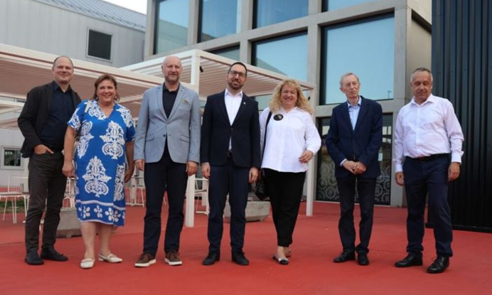 Fotografija prikazuje sedam osoba koje stoje na crvenom podu ispred moderne zgrade s velikim staklenim prozorima. Svi su odjeveni poslovno—sakoa, košulje, hlače i elegantne haljine dominiraju—što upućuje na neki službeni događaj, poput poslovnog sastanka, konferencije ili svečanosti. U pozadini se vidi bijela pergola, a ispod nje nekoliko stolova i stolica, što daje prostoriji otvoreni, ali organizirani karakter. Sve zajedno odiše profesionalnošću i suvremenim urbanim stilom.