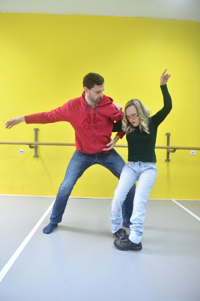 Muškarac u crvenoj dukserici i trapericama drži ženu s dugom plavom kosom i naočalama, koja je odjevena u crnu majicu i svijetle traperice. Zajedno izvode plesnu figuru u dvorani s jarko žutim zidom i baletnim šipkama.