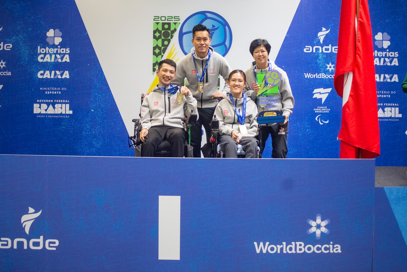 Fotografija prikazuje tri čovjeka u invalidskim kolicima s medaljama oko vrata. Oni stoje na mjestu s brojem jedan što sugerira da su osvojili prvo mjesto.