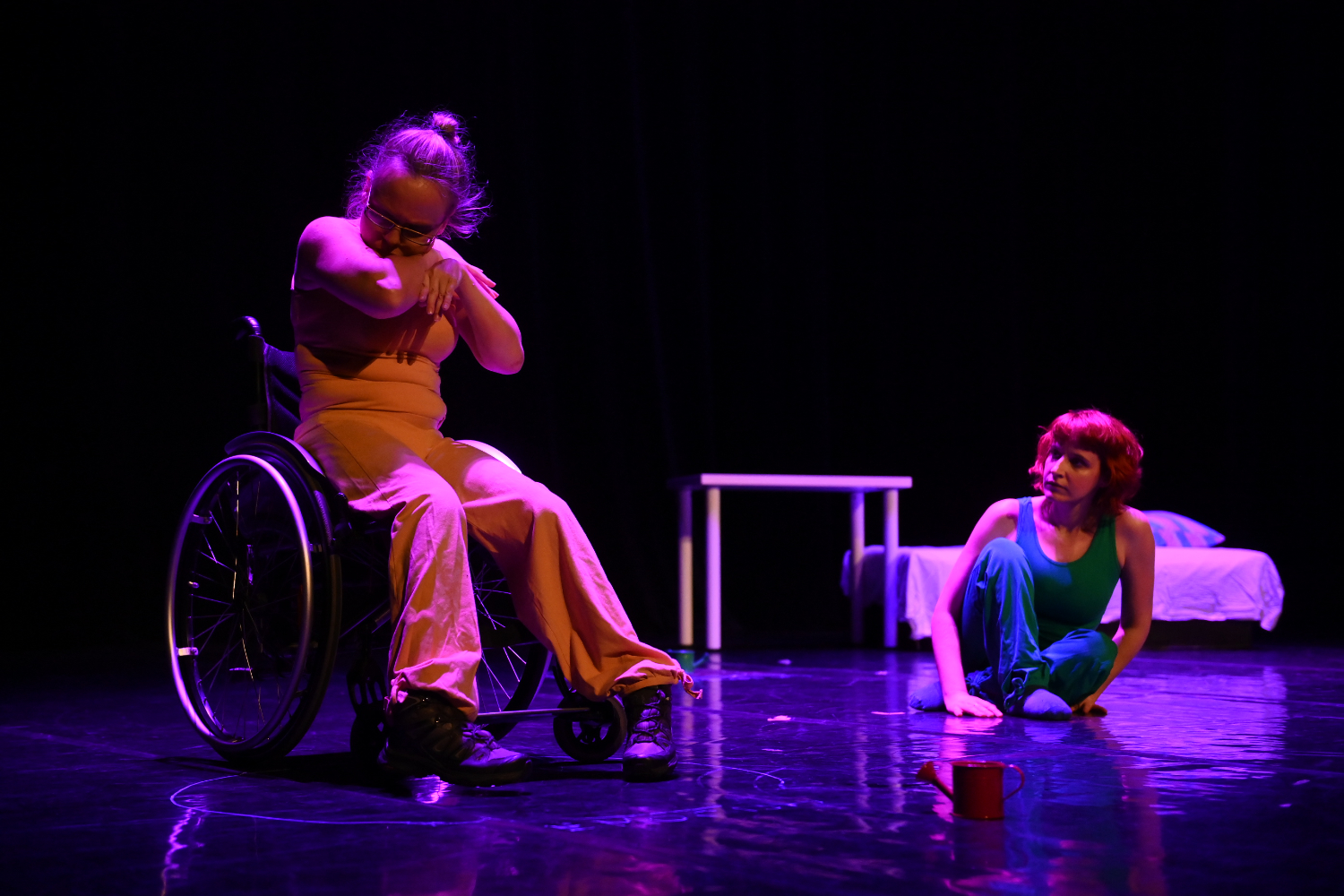 Žena u invalidskim kolicima odjevena u narančasti kostim izvodi pokret rukama, dok druga žena s crvenom kosom u zelenom kostimu sjedi na podu i promatra je. Na pozornici su stol, krevet i crvena zalijevača, uz umjetničko svjetlo koje stvara dramatičnu atmosferu.