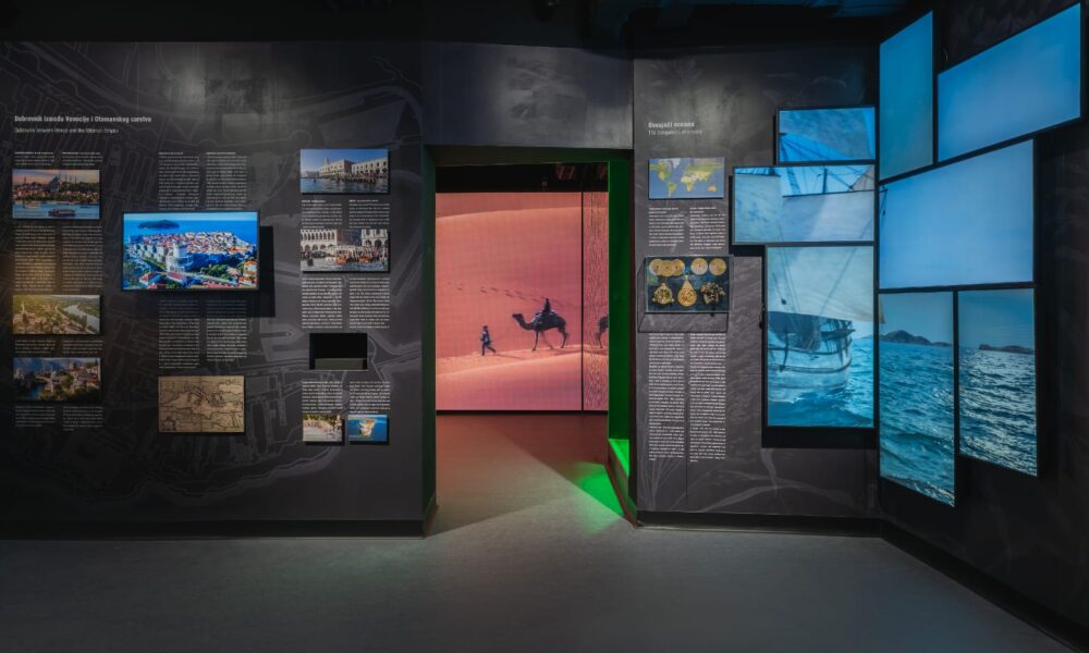 Unutrašnjost modernog muzeja ili izložbenog prostora s tamnim zidovima na kojima su postavljeni tekstovi, povijesne fotografije, zemljovidi i interaktivni ekrani. Središnji prolaz vodi prema svijetloj digitalnoj projekciji pješčane pustinje s osobom i devom. Desno su ekrani s prikazima mora i jedrenjaka, dok se lijevo prikazuju prizori Dubrovnika, Venecije i Osmanskog carstva.