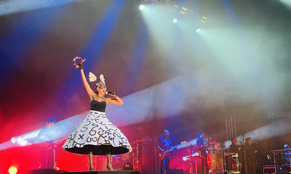 Pjevačica na pozornici nosi crno-bijelu haljinu s velikim slovima i glavu ukrašenu krilima, držeći tamburin visoko iznad glave dok pjeva u mikrofon. Iza nje svira bend, a pozornicu obasjavaju jarka crvena, plava i bijela svjetla.