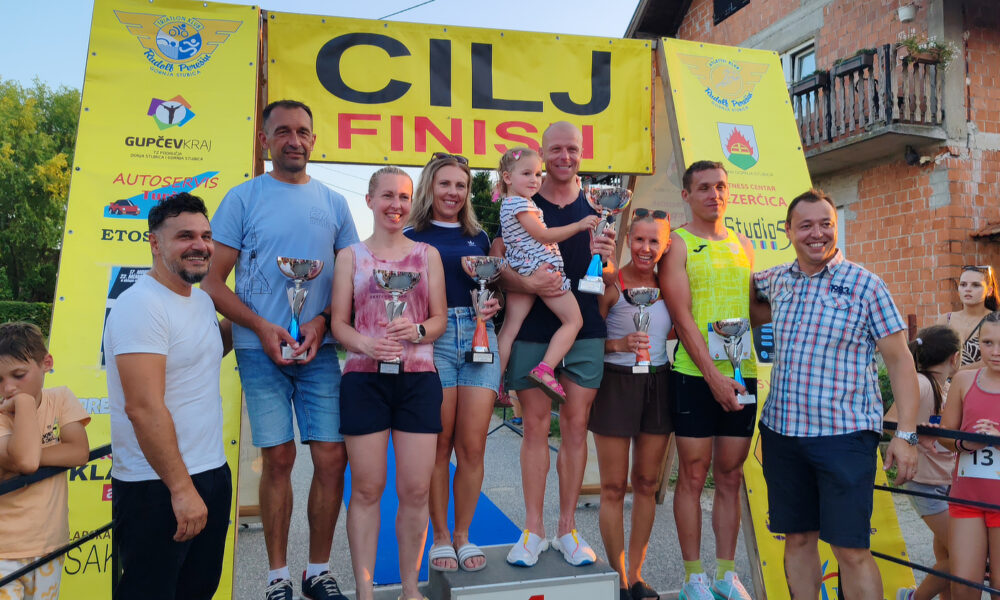 Skupina muškaraca i žena stoji na postolju ispred natpisa „CILJ/FINISH“, drže pehare nasmiješeni, dok ih okružuje publika. Jedan muškarac drži malo dijete u naručju.