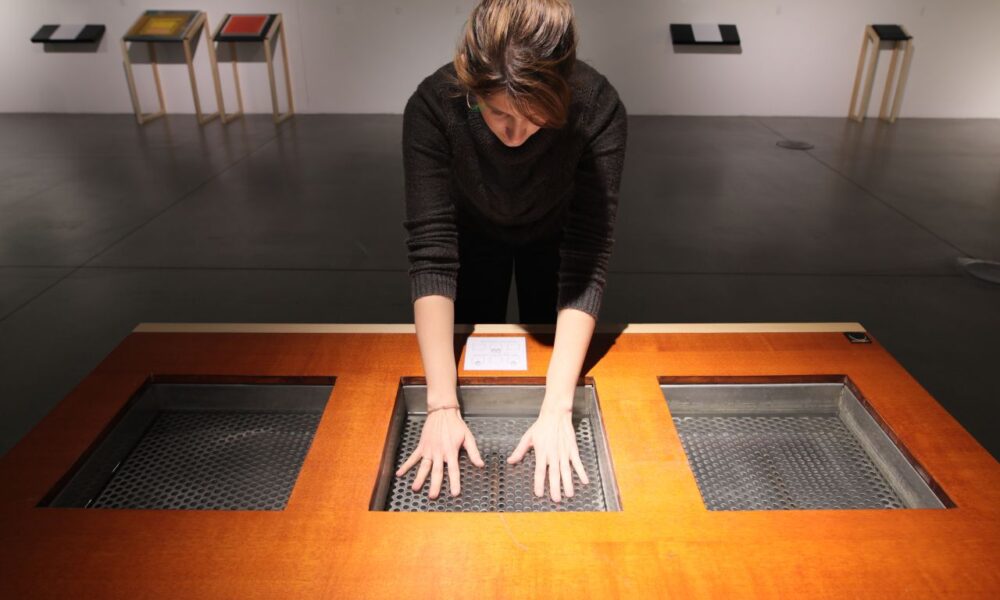 Žena stoji nad drvenim stolom s tri kvadratna otvora, pružajući ruke prema unutra kako bi dodirnula unutrašnje površine s rupičastom teksturom.