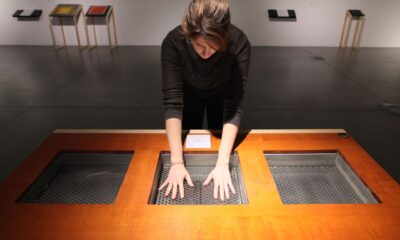 Žena stoji nad drvenim stolom s tri kvadratna otvora, pružajući ruke prema unutra kako bi dodirnula unutrašnje površine s rupičastom teksturom.