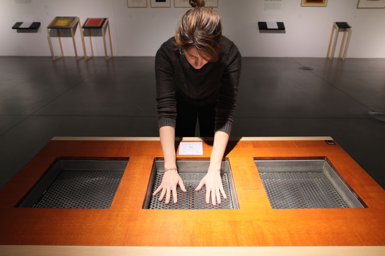 Žena stoji nad drvenim stolom s tri kvadratna otvora, pružajući ruke prema unutra kako bi dodirnula unutrašnje površine s rupičastom teksturom.