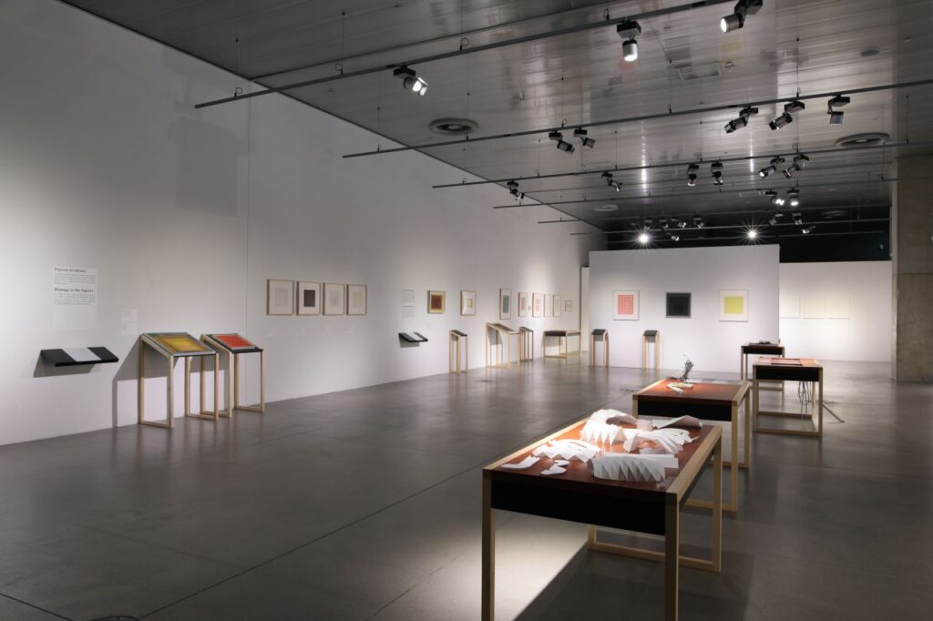 Prostrana galerija s bijelim zidovima na kojima vise geometrijske umjetnine i tekstualne ploče, dok su u prostoru raspoređeni drveni stolovi s izloženim radovima i maketama pod staklom.