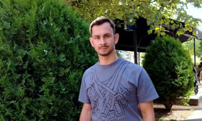 Mladić kratke kose i brade stoji ispred zelenog grmlja i kafića na otvorenom. Odjeven je u sivu majicu s geometrijskim uzorkom i gleda ravno u kameru.