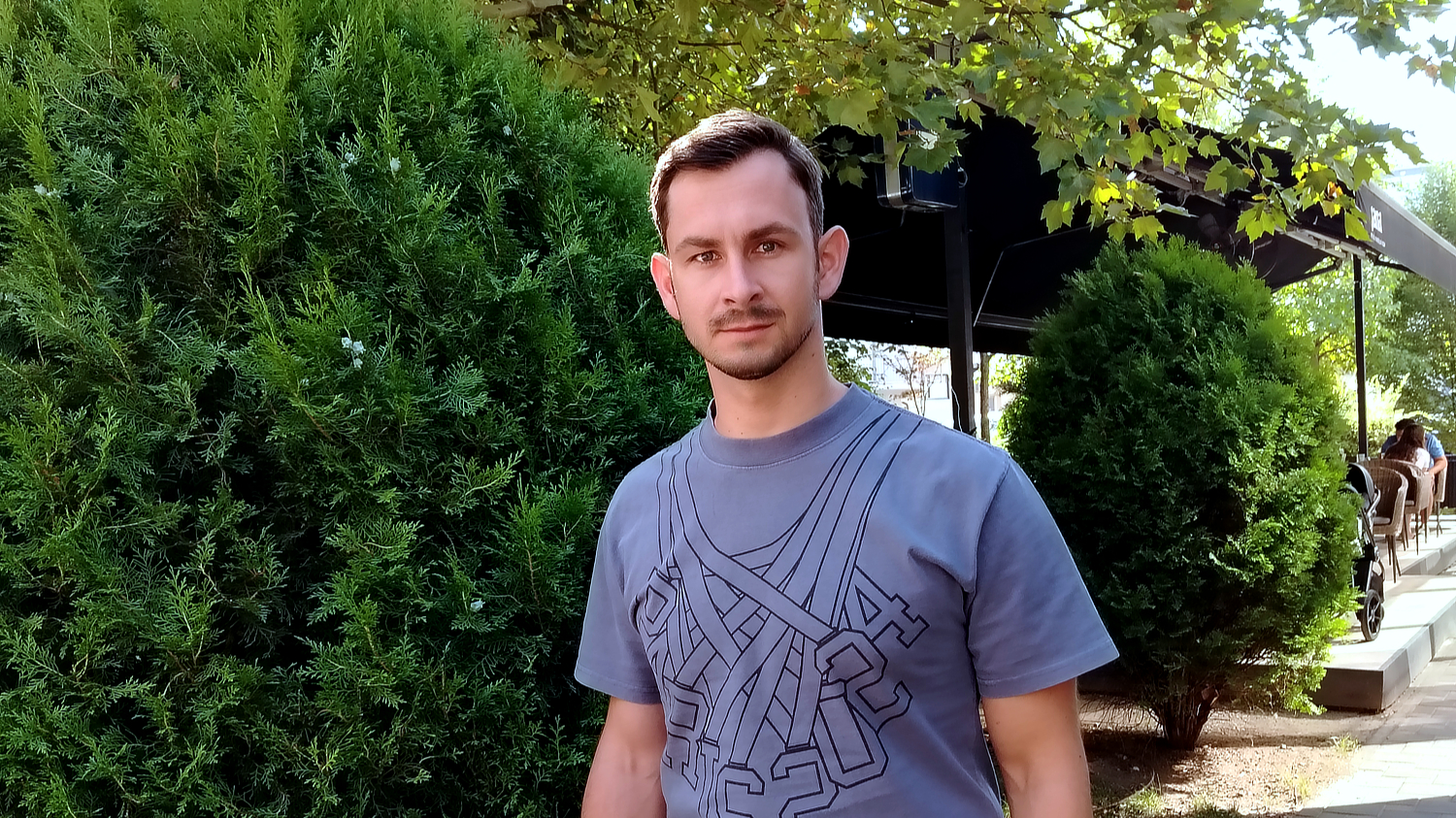 Mladić kratke kose i brade stoji ispred zelenog grmlja i kafića na otvorenom. Odjeven je u sivu majicu s geometrijskim uzorkom i gleda ravno u kameru.