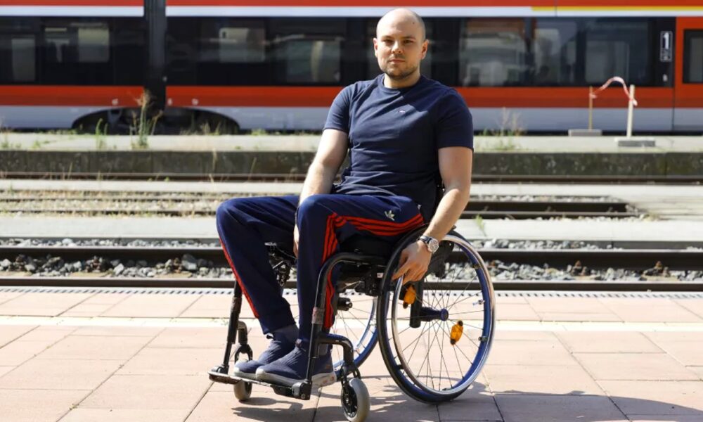 Na slici je prikazana osoba u invalidskim kolicima koja sjedi na pločniku uz željezničku prugu. Odjevena je u tamnoplavu majicu kratkih rukava i tamnoplave sportske hlače s crvenim prugama. U pozadini se nalazi crveno-sivi vlak koji stoji na stanici.