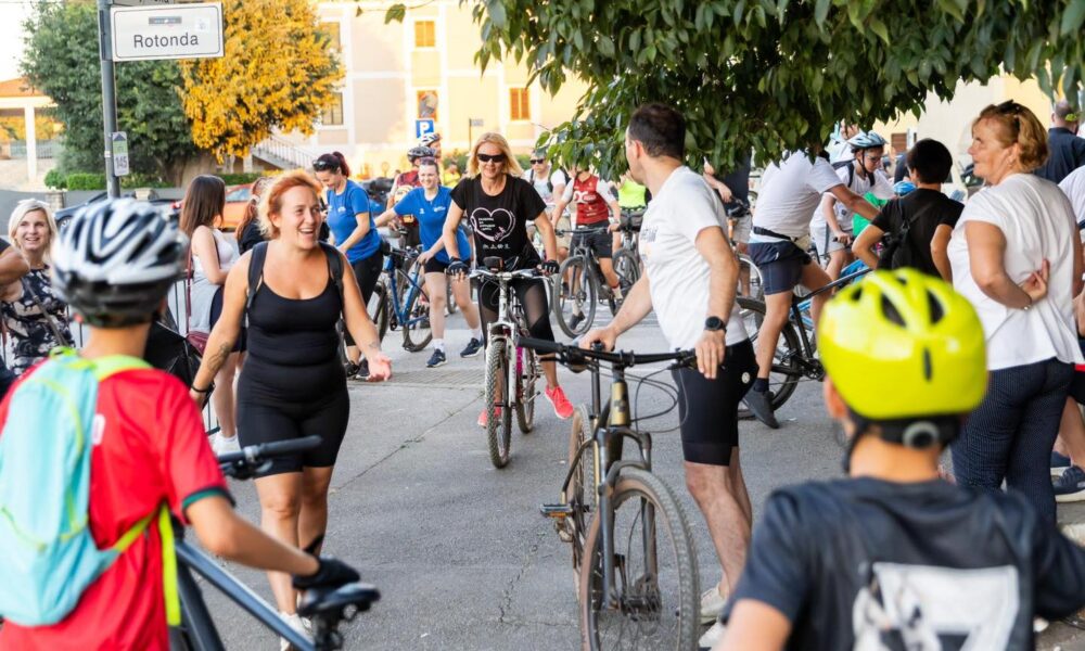 Fotografija prikazuje više ljudi na biciklima te neke ljude koji stoje.