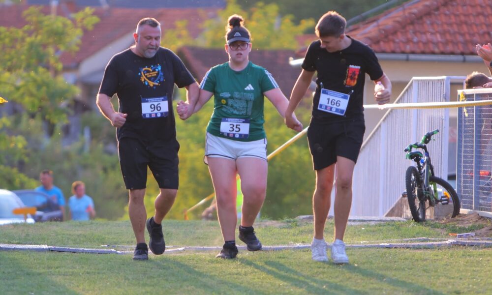 Fotografija prikazuje troje trkača kako prelaze liniju cilja držeći se za ruke, Jedno od to troje trkača ima Down sindrom.