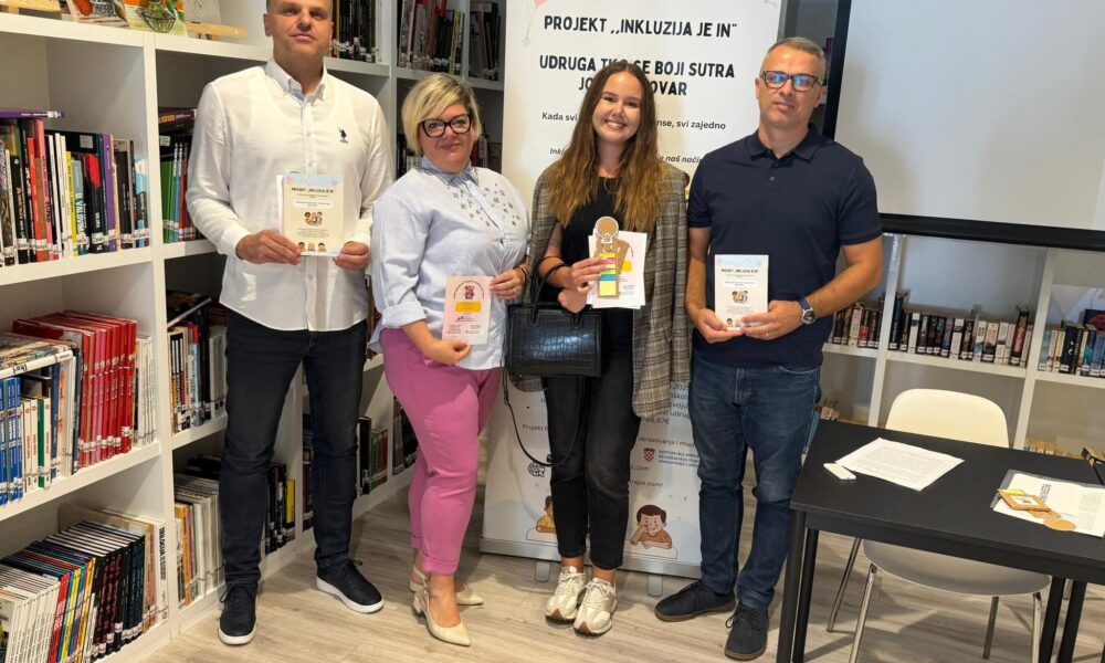 Na fotografiji se nalaze četiri odrasle osobe koje stoje u knjižnici ili knjižari, svaka drži knjigu ili knjižicu u rukama. Iza njih je istaknut banner s tekstom na hrvatskom jeziku: "PROJEKT 'INKLUZIJA JE IN' UDRUGA 'JEDNAKI U BOJI SUTRA' KUTAK ZA INKLUZIJU – Mjesto gdje se različitost slavi, a ne zaboravlja"
