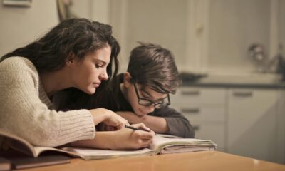 Na fotografiji su dvije osobe koje sjede za stolom u kućnom ambijentu — najvjerojatnije kuhinji ili blagovaonici, s obzirom na vidljive ormariće i radnu površinu u pozadini. Atmosfera je topla i domaća. Osoba s lijeve strane pokazuje nešto u otvorenoj bilježnici ili udžbeniku, dok osoba s desne strane piše ili crta, koncentrirano prateći upute. Izraz lica obiju osoba odaje ozbiljnost i fokusiranost, što sugerira da se radi o učenju, možda domaćoj zadaći ili instrukcijama. Ova scena lijepo prikazuje trenutak podrške i zajedničkog rada — mogla bi biti roditelj koji pomaže djetetu, stariji brat ili sestra, ili čak privatni instruktor. U svakom slučaju, fotografija prenosi poruku o važnosti obrazovanja, strpljenja i međusobne pomoći.