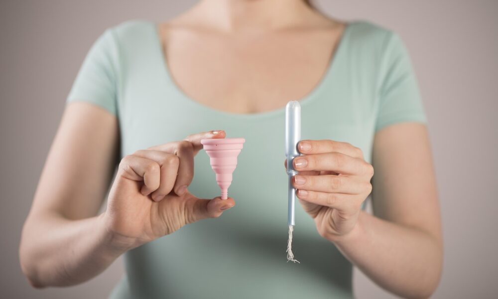 Na fotografiji je prikazana osoba koja u lijevoj ruci drži ružičastu menstrualnu čašicu, a u desnoj plavi tampon s bijelim koncem. Osoba nosi svijetlozelenu majicu. Ova slika jasno ilustrira dvije različite vrste menstrualnih proizvoda, što može biti korisno za edukaciju i usporedbu opcija za menstrualnu higijenu.
