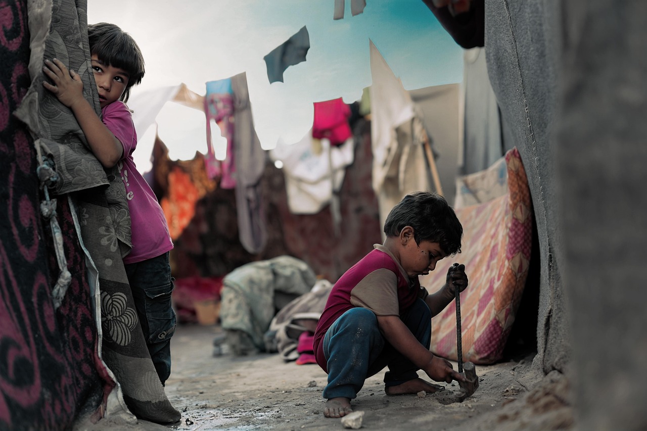 Na fotografiji se nalaze dvoje djece u siromašnom, improviziranom okruženju. Jedno dijete, odjeveno u rozu majicu, skriva se iza izblijedjelog tepiha i s oprezom promatra u daljinu. Drugo dijete, boso i u odjeći koja je prljava od zemlje, sjedi na tlu i nešto kucka ili pokušava razbiti pomoću metalnog predmeta. Oko njih vise oprani komadi odjeće i razne krpe, a u pozadini se vidi skromno i improvizirano sklonište od raznobojnih tkanina i madraca. Cijela scena odiše osjećajem siromaštva, nesigurnosti i borbe za osnovne uvjete života.