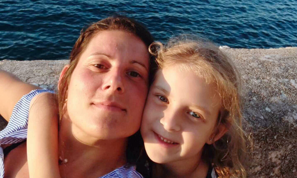 Žena i djevojčica poziraju zagrljene za selfie uz obalu mora. U pozadini se vidi plavetnilo morske površine, a obje se blago smiješe u kameru, stvarajući topao i blizak obiteljski trenutak.