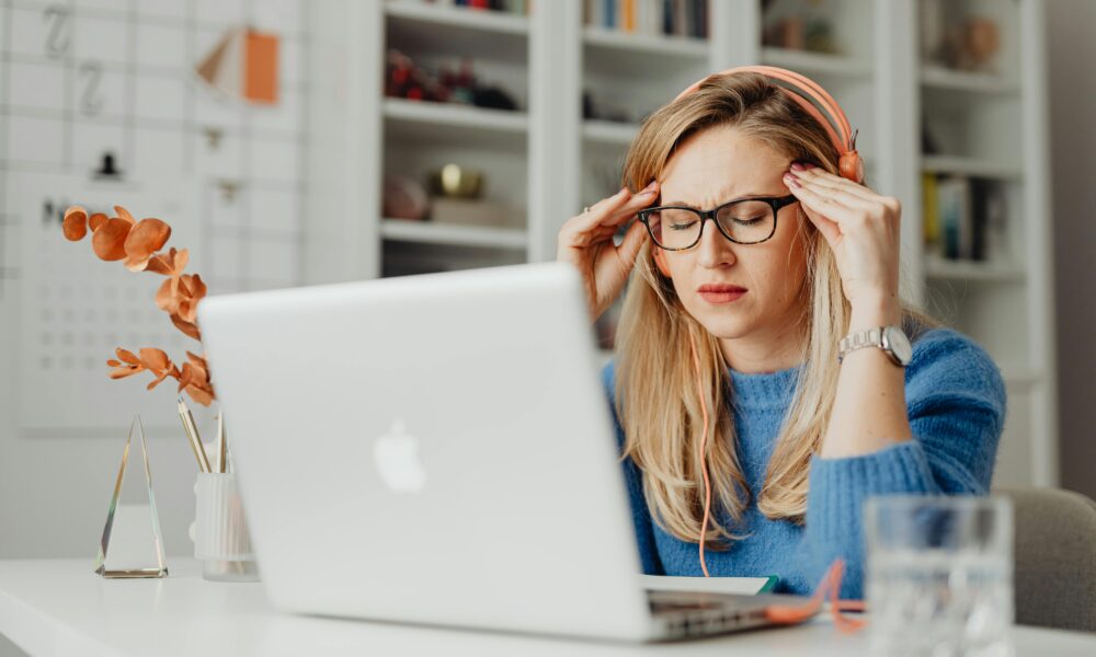 Na fotografiji se nalazi osoba koja sjedi za radnim stolom ispred otvorenog Apple MacBook laptopa. Osoba nosi plavi pulover i narančaste slušalice, a djeluje kao da osjeća stres ili nelagodu — ruke su joj položene na sljepoočnice, a oči zatvorene.