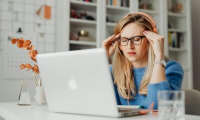 Na fotografiji se nalazi osoba koja sjedi za radnim stolom ispred otvorenog Apple MacBook laptopa. Osoba nosi plavi pulover i narančaste slušalice, a djeluje kao da osjeća stres ili nelagodu — ruke su joj položene na sljepoočnice, a oči zatvorene.