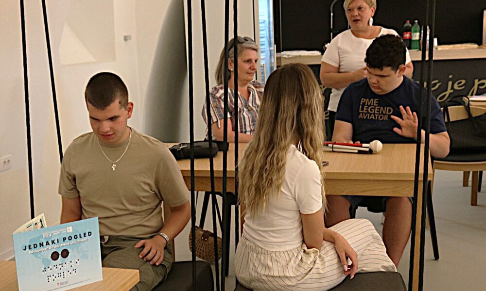 Fotografija prikazuje nekoliko ljudi koji sjede u klupama. Ispred jednog dečka je brošura 'Jednaki pogled'.