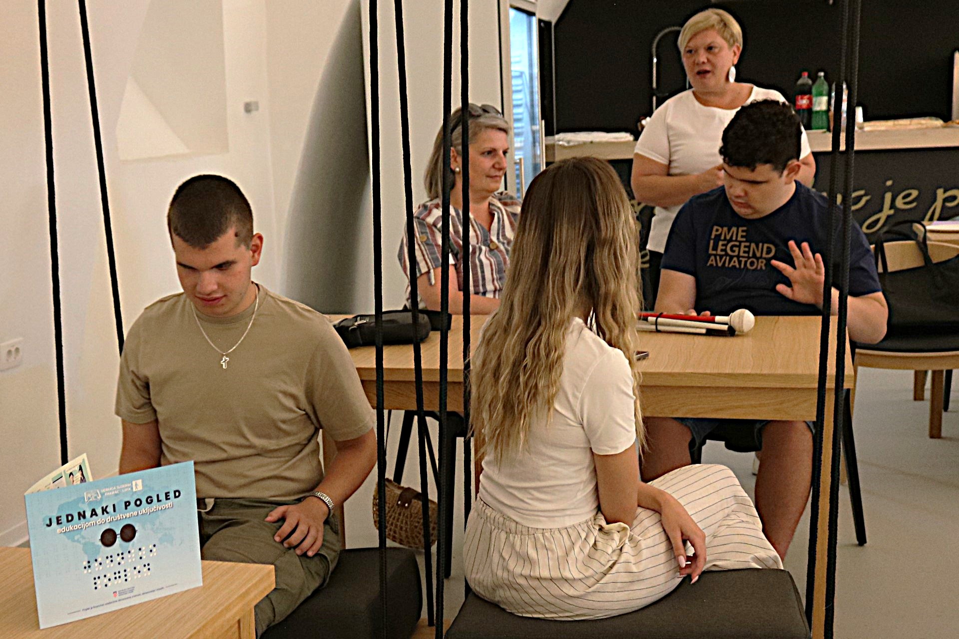 Fotografija prikazuje nekoliko ljudi koji sjede u klupama. Ispred jednog dečka je brošura 'Jednaki pogled'.