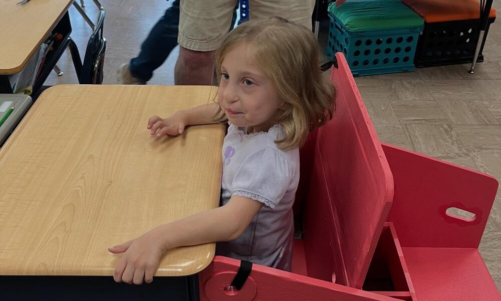 Fotografija prikazuje curicu s invaliditetom kako sjedi za prilagođenom stolom.