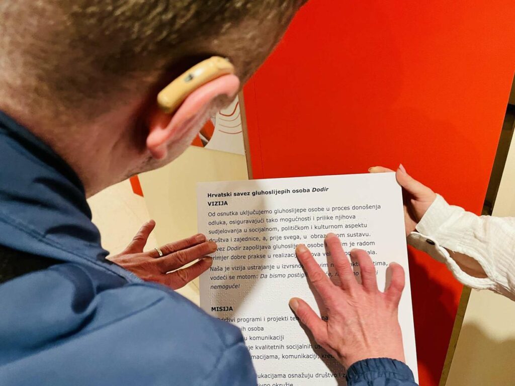 Osoba sa slušnim aparatom i druga osoba zajedno drže ploču s tekstom na hrvatskom jeziku o viziji i misiji Hrvatskog saveza gluhoslijepih osoba Dodir. Ruke prelaze preko teksta, naglašavajući važnost pristupačnosti i uključivanja.