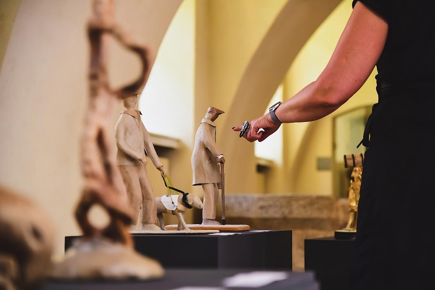 Ruka osobe pokazuje na male figurativne skulpture postavljene na crnim postoljima u izložbenom prostoru s kamenim svodovima.