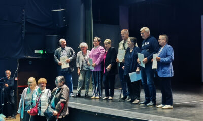 Skupina starijih osoba stoji na pozornici i drži priznanja, dok nekoliko ljudi u publici snima i promatra događaj.