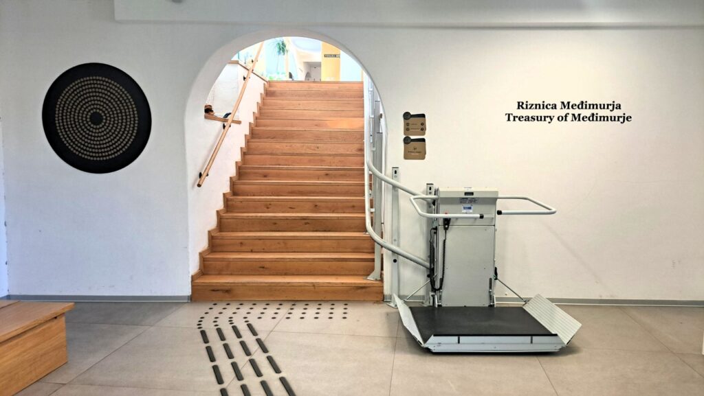 Ulazni prostor muzeja s drvenim stubištem koje vodi na kat. S desne strane nalazi se platformno dizalo za osobe s invaliditetom, a na podu su taktilne oznake za slijepe i slabovidne osobe. Na zidu piše „Riznica Međimurja / Treasury of Međimurje“.