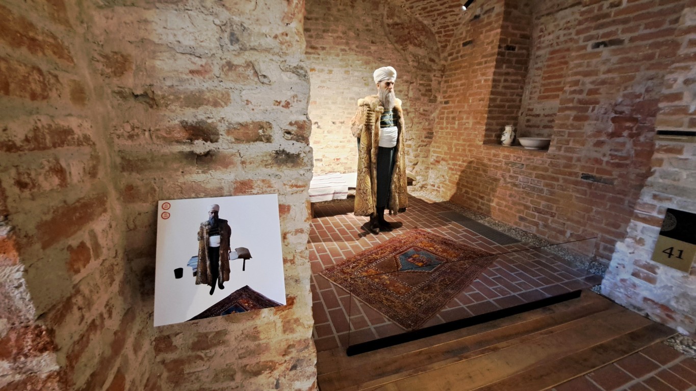 Unutrašnjost muzeja s izloženom figurom muškarca u povijesnoj odjeći s turbanom i bogato ukrašenim kaputom. Figura je smještena u nišu od crvene opeke, a ispred se nalazi šareni sag. Na zidu je ploča s fotografijom iste figure za dodatno objašnjenje izložbe.